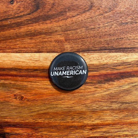1.5" Round black button with 'MAKE RACISM UNAMERICAN' text on a wooden surface.
