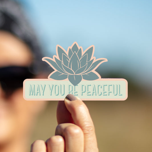 Hand holding a card with a lotus flower design and 'May You Be Peaceful' text against a blurred natural background.