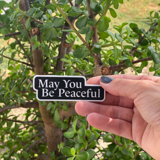 Hand holding a black and white metta inspired Buddhist mantra sticker that reads 'May You Be Peaceful' in front of green foliage.