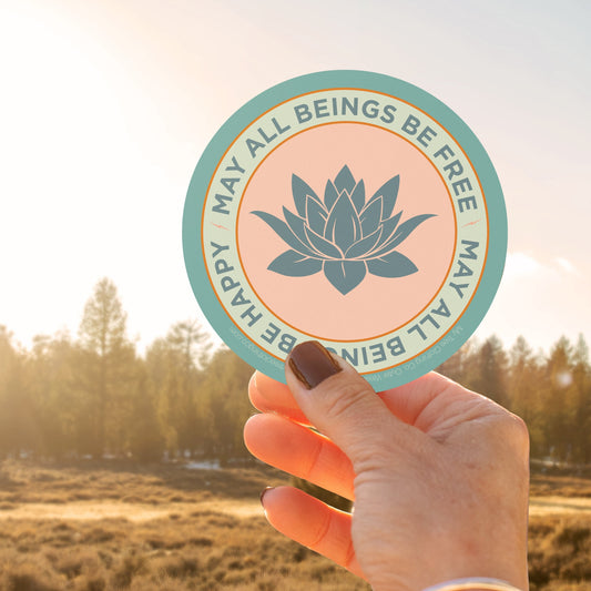 Hand holding a circular sticker with a lotus flower design and text that reads, may all beings be free may all beings be happy, against a sunset background.