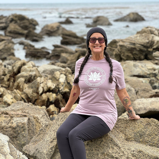 Person sitting on rocks by the ocean wearing a light purple t-shirt with a white lotus in the center and the words, may you be peaceful may you be happy may you be free.