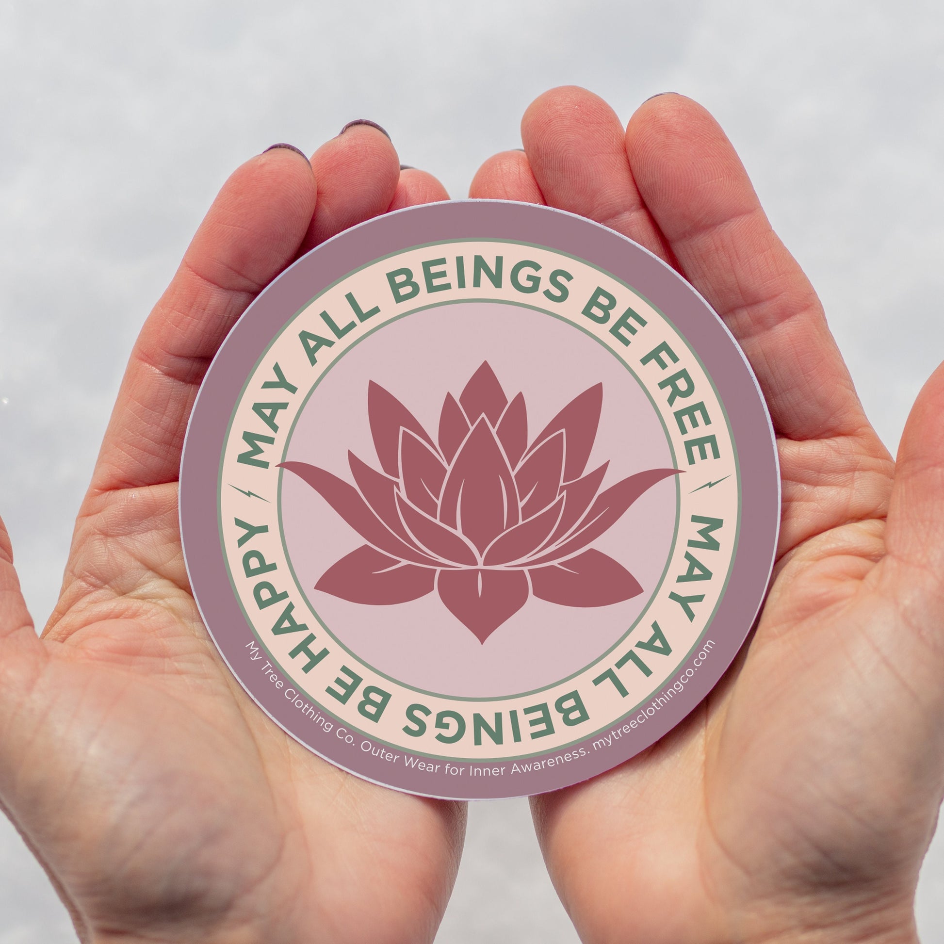 4" Circular sticker with lotus flower design and the text, MAY ALL BEINGS BE FREE MAY ALL BEINGS BE HAPPY held between two hands against a white background.
