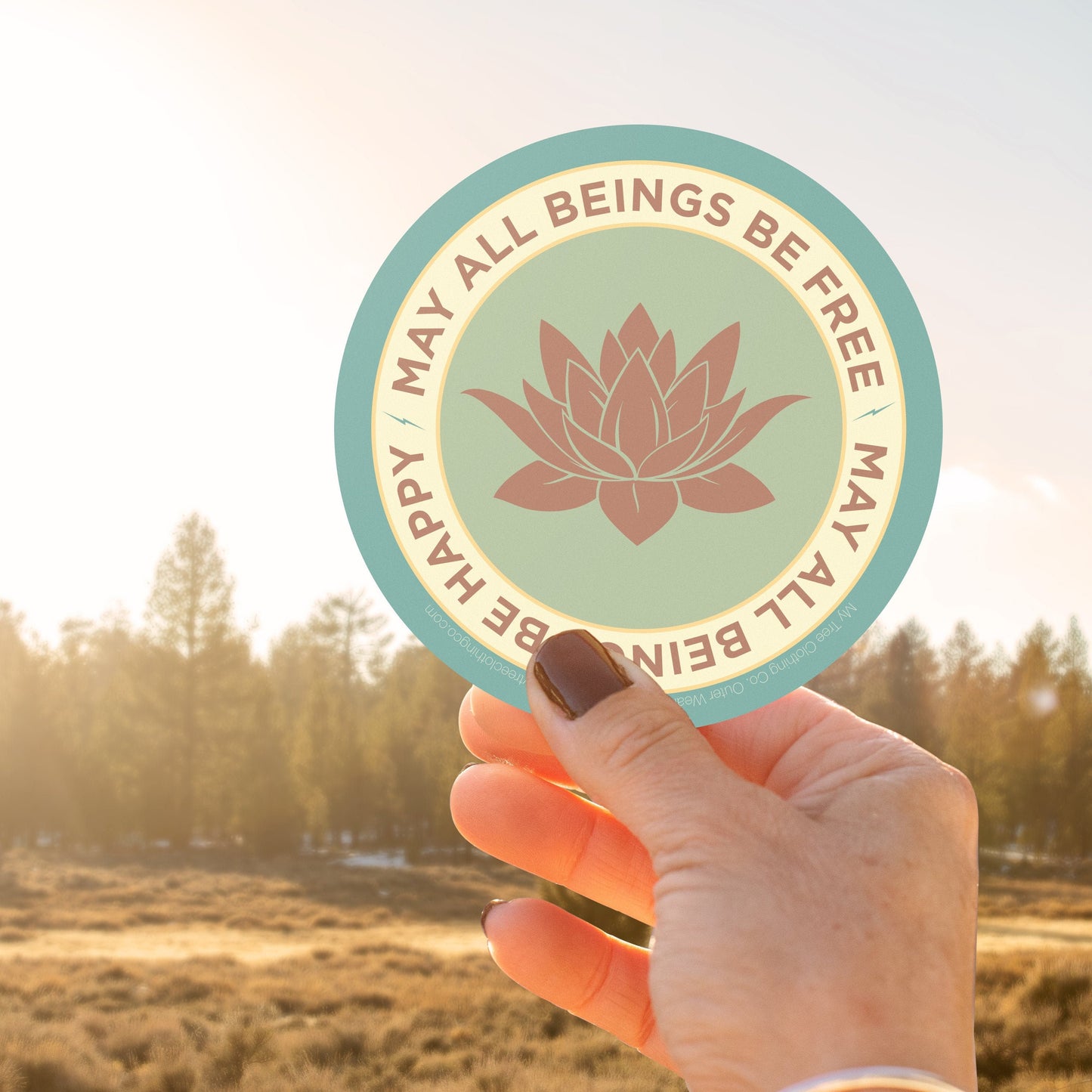 Hand holding a circular sticker with a lotus flower design and text, MAY ALL BEINGS BE FREE MAY ALL BEINGS BE HAPPY, against a natural background.