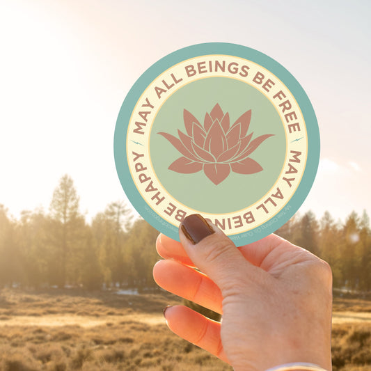 Hand holding a circular sticker with a lotus flower design and text, MAY ALL BEINGS BE FREE MAY ALL BEINGS BE HAPPY, against a natural background.