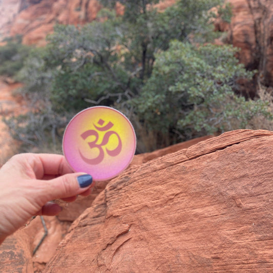 Hand holding a pink and gold 3" circular Om symbol sticker against a natural background.