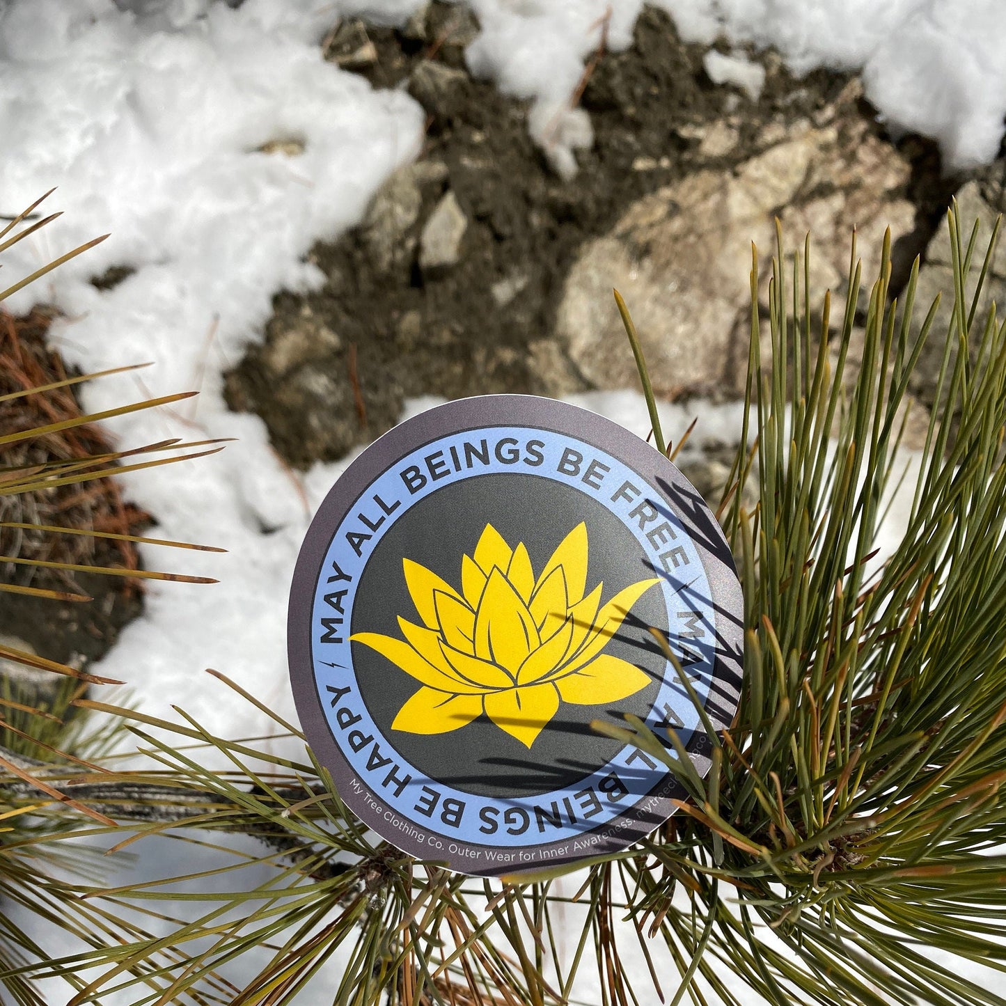 Round sticker with yellow buddhist lotus flower design and text, may all beings be free may all beings be happy, on a pine branch with snow in the background