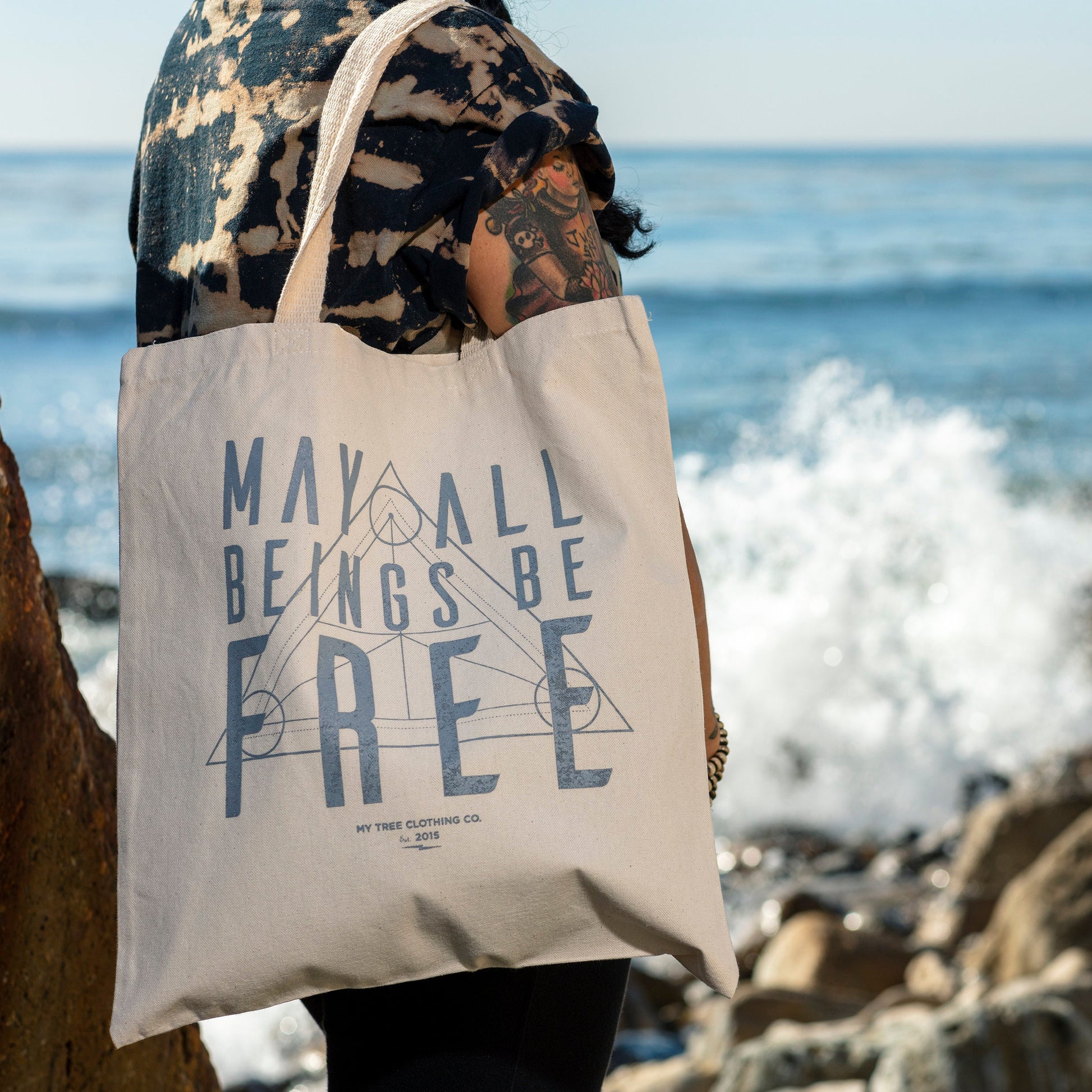 Person holding a tote bag with the text, MAY ALL BEINGS BE FREE, with a sacred geometry design with ocean waves in the background.
