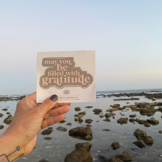 Hand holding a sticker with 'may you be filled with gratitude' text against a coastal background.