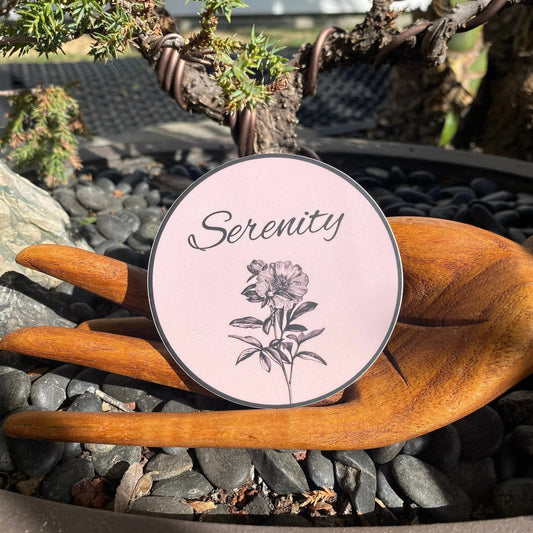 Round pink 3" round sticker with 'Serenity' and floral design on a wooden surface outdoors.