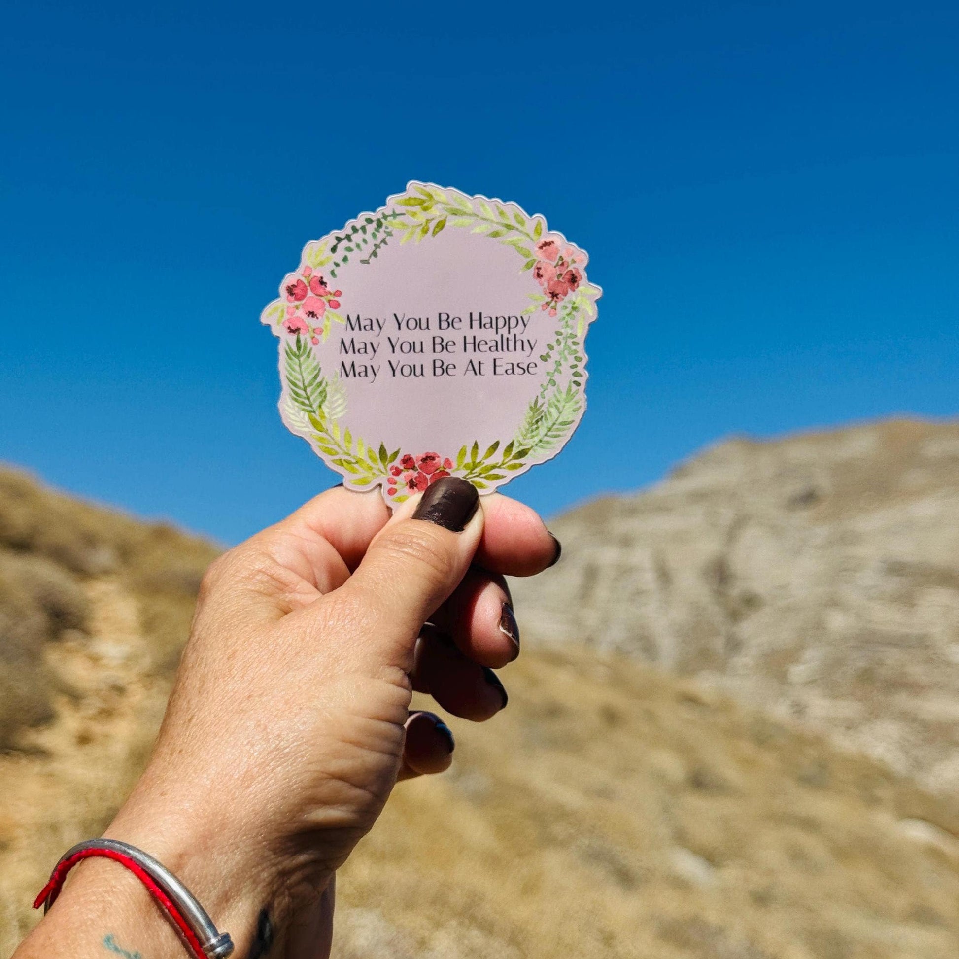 Hand holding a circular sticker with floral design and text, may you be happy, may you be healthy, may you be at ease, against a blue sky and mountain background
