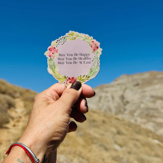 Hand holding a circular sticker with floral design and text, may you be happy, may you be healthy, may you be at ease, against a blue sky and mountain background