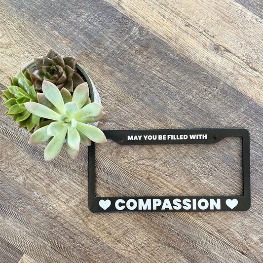 Black license plate frame with 'May you be filled with compassion' text on a wooden surface with succulents.