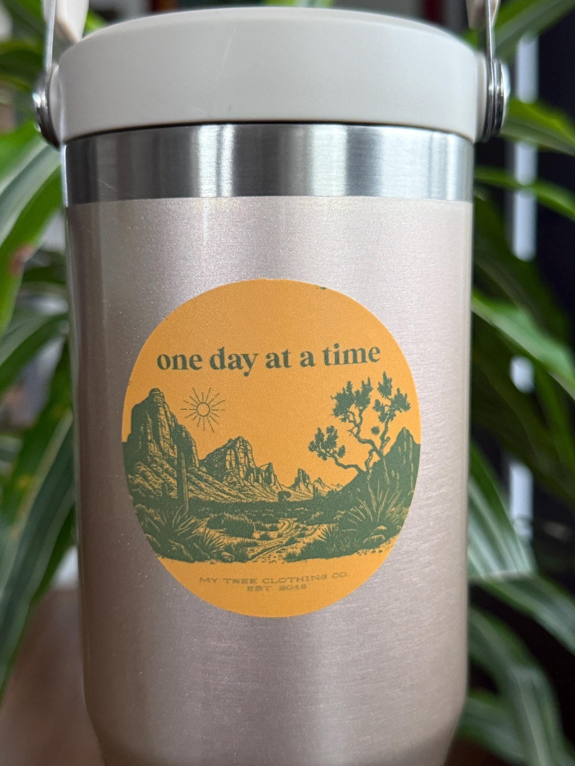 Stainless steel mug with 'one day at a time' logo and mountain design, held against a blurred green background.