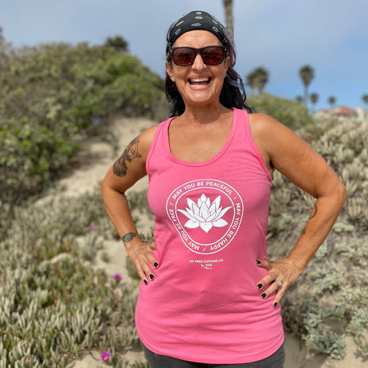 Woman wearing a pink tank top with a white lotus flower design and text that reads, 'may you be peaceful may you be happy may you be free' standing outdoors with greenery in the background.