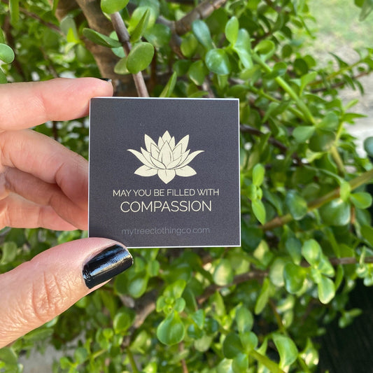 Hand holding a 2" square lotus flower sticker with text 'May you be filled with compassion' against a green leafy background.