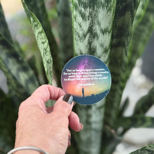 Hand holding a round sticker with a prayer by shantideva in front of a plant