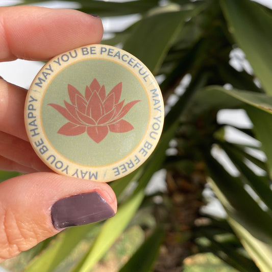 Round button with lotus flower design held by a hand against a green leafy background. Text on button from metta prayer, may you be peaceful may you be free may you be happy.