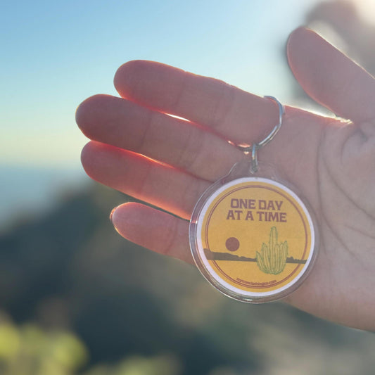 Hand holding a keychain with 'One Day at a Time' text and cactus illustration against a blurred natural background.