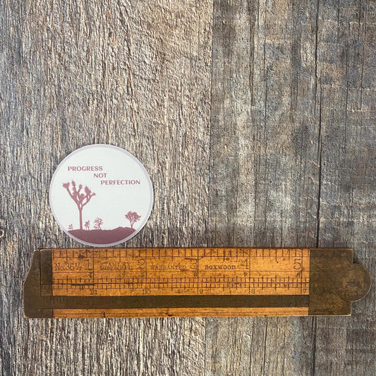 Wooden ruler with a circular 12 step recovery sticker with the text, Progress Not Perfection, on a wooden surface.