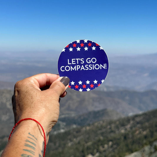 Hand holding a patriotic red, white, and blue, 'Let's Go Compassion!' sticker with a mountainous landscape in the background. Real patriots support kindness, inclusion, and diversity!