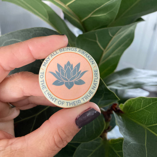 Hand holding a 1.5" round lotus design button with the text, may all beings awaken to the light of their true nature, against a green leafy background.
