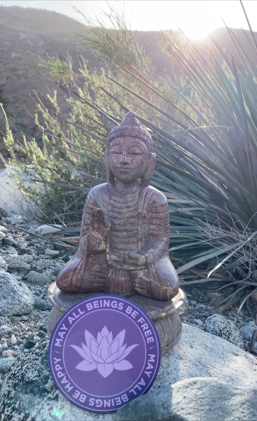 Buddha statue with a purple magnet with lotus design and text, may all beings be free may all beings be happy, in a natural setting.