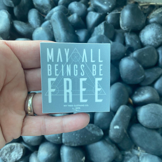 Hand holding a 2" square grey sticker with 'May all beings be free' text with sacred geometry design, held in front of rocks.