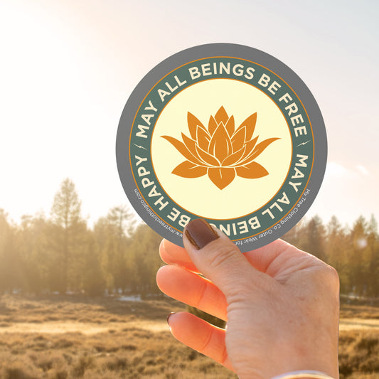 Hand holding a 4" circular sticker with a saffron colored lotus flower design and Mettā prayer, against a blurred natural background.