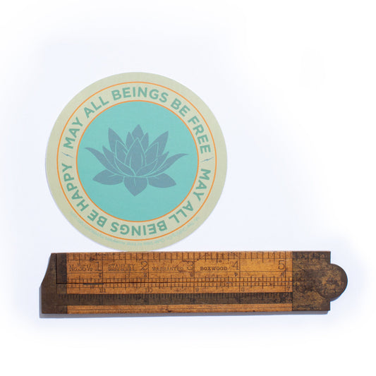 Round teal sticker with lotus flower design and text,  may all beings be free may all beings be happy, on a wooden ruler stand.