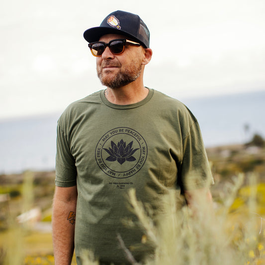 Man wearing a green t-shirt with a lotus in the center that says, may you be peaceful, happy, free. They're standing in a natural setting.