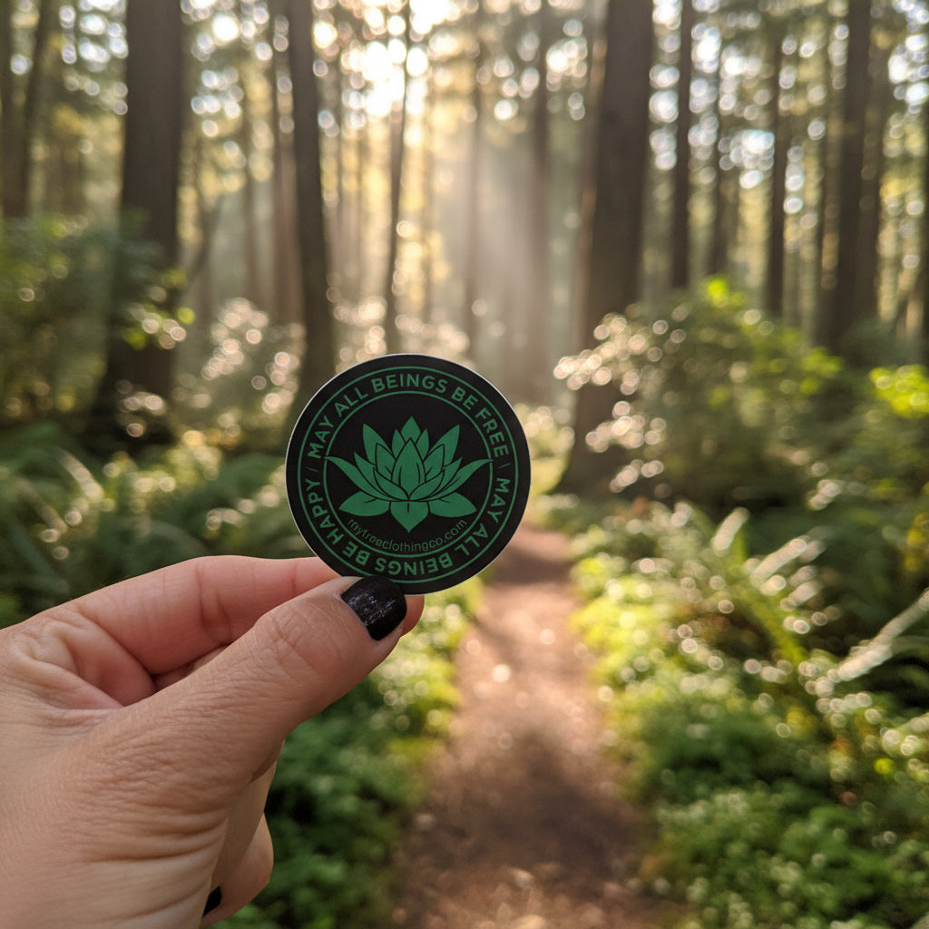 Two inch round green and black lotus sticker with the words, MAY ALL BEINGS BE FREE-MAY ALL BEINGS BE HAPPY. It's being measured by an antique wooden ruler. Being held in a hand with a nature scene in the bakground.