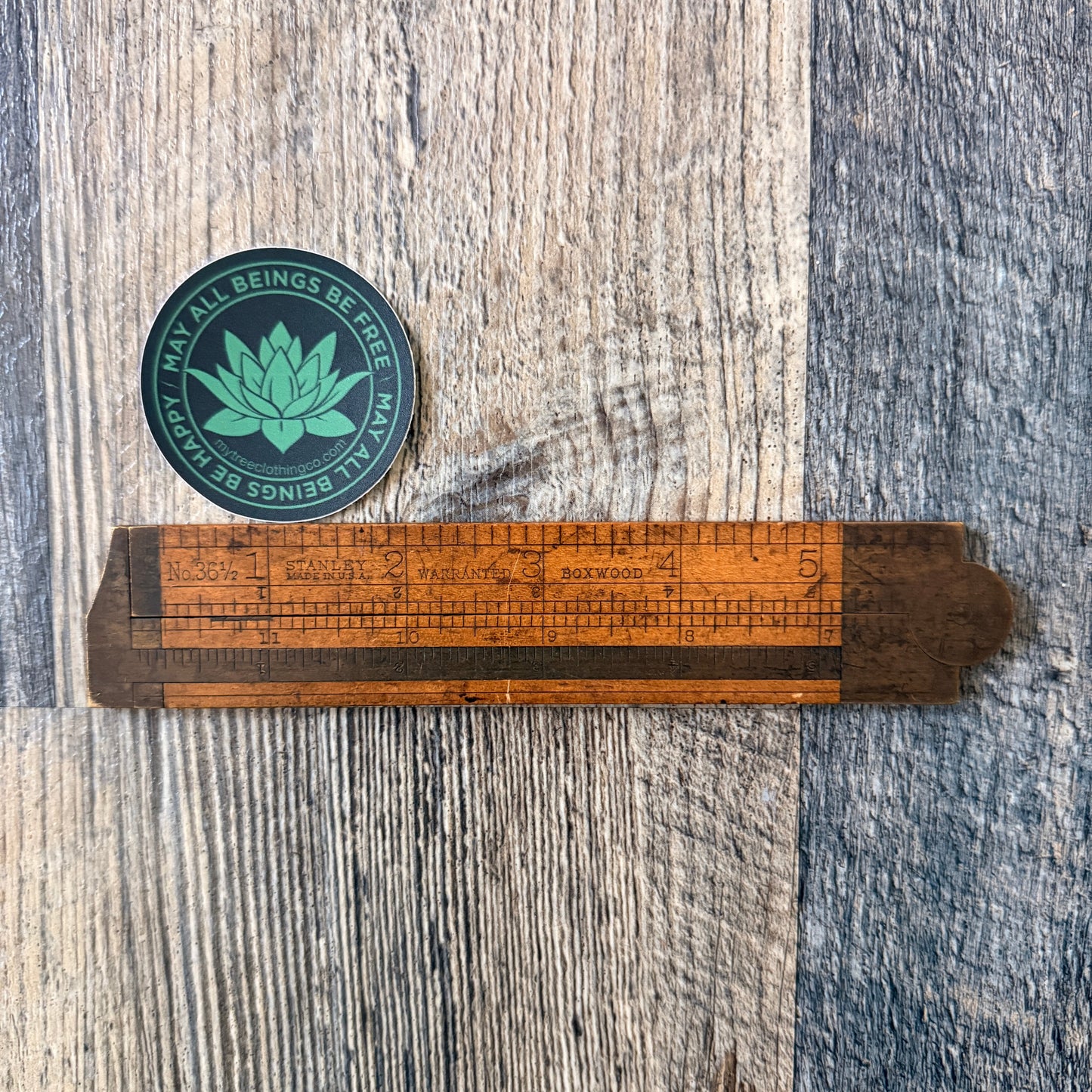 Two inch round green and black lotus sticker with the words, MAY ALL BEINGS BE FREE-MAY ALL BEINGS BE HAPPY. It's being measured by an antique wooden ruler.