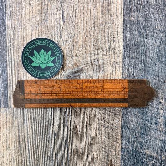 Two inch round green and black lotus sticker with the words, MAY ALL BEINGS BE FREE-MAY ALL BEINGS BE HAPPY. It's being measured by an antique wooden ruler.