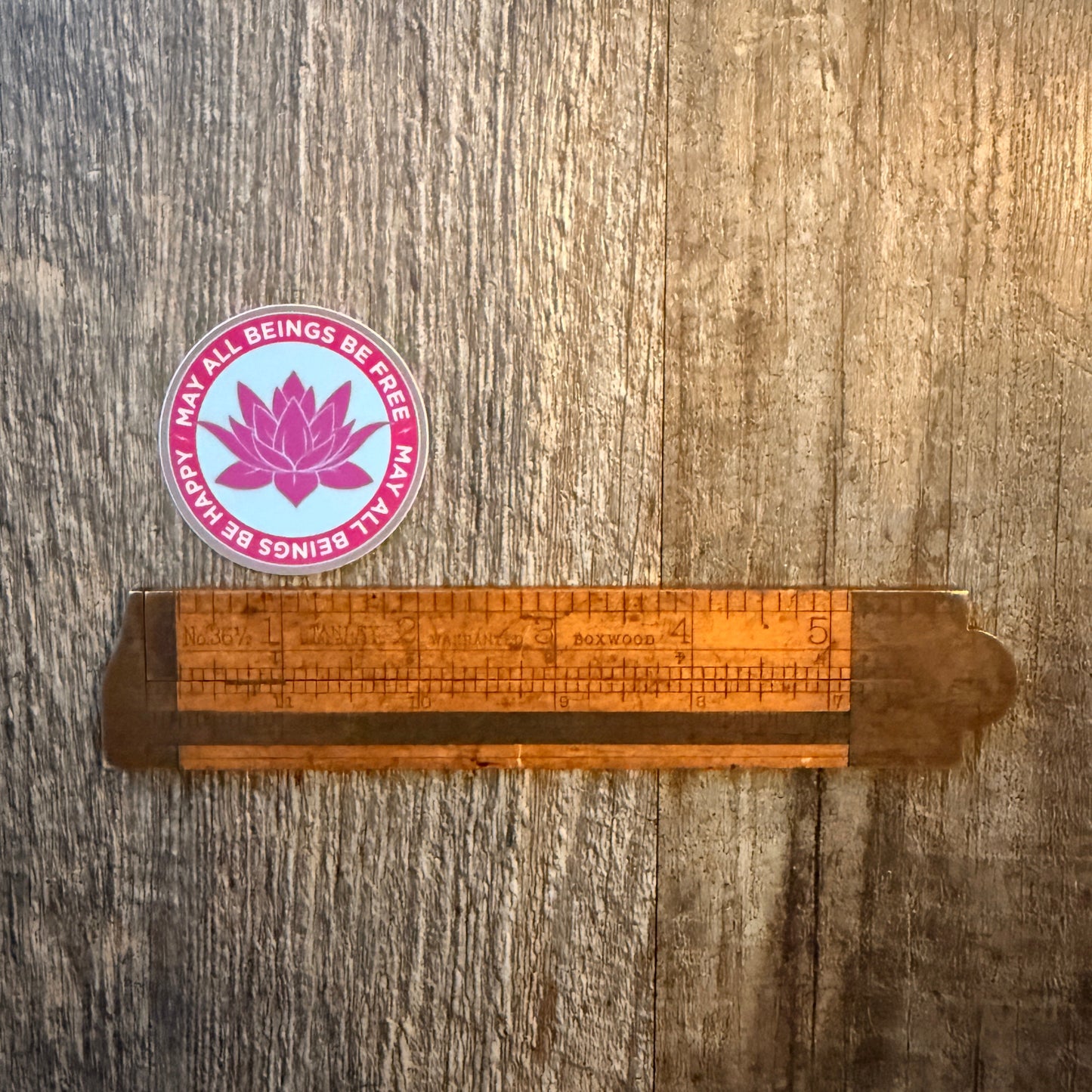 Wooden ruler on a wooden surface with a pink and purple sticker featuring a lotus flower design and the Buddhist mantra, may all beings be free-may all beings be happy.
