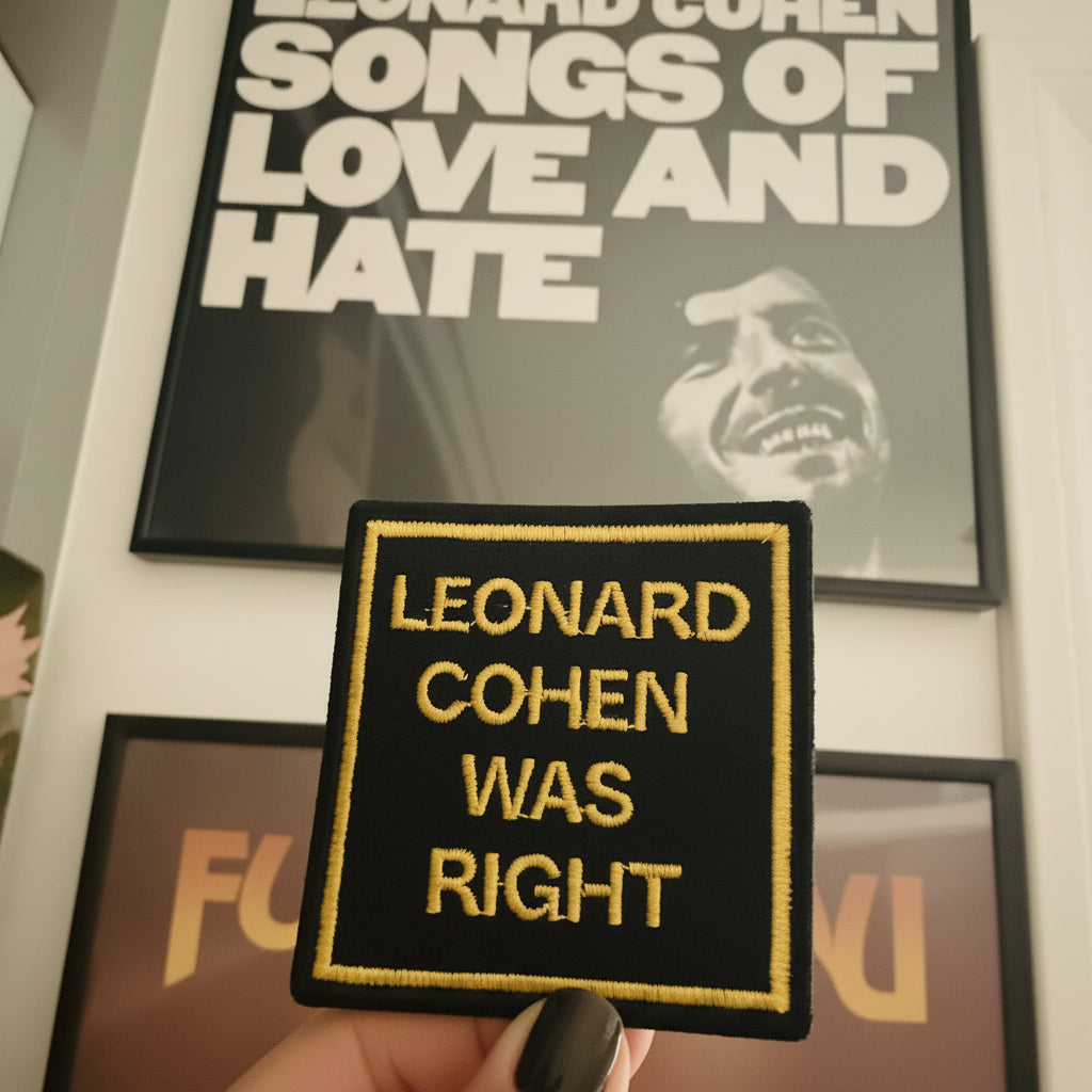 A hand with black nail polish holds a rectangular embroidered patch that reads 'LEONARD COHEN WAS RIGHT' in yellow text on a black background with a yellow border. Behind it are two framed Leonard Cohen posters on a wall.