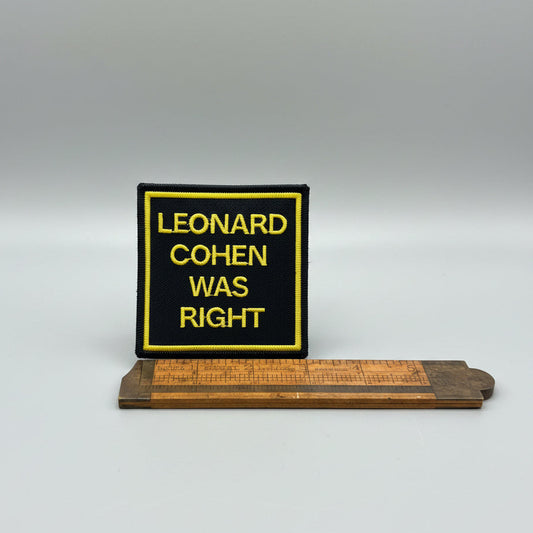 A 3" square black embroidered patch with yellow border and yellow text reading 'LEONARD COHEN WAS RIGHT' is placed on a wooden surface above a wooden ruler for scale.