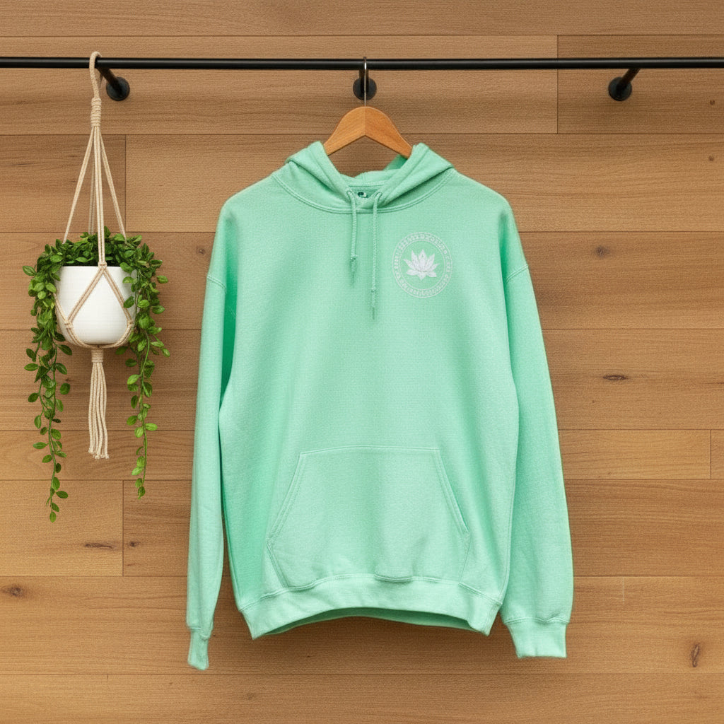 Mint green hoodie with a lotus and the text, may you be happy may you be free may you be peaceful on a wooden hanger against a wooden wall and black rod.