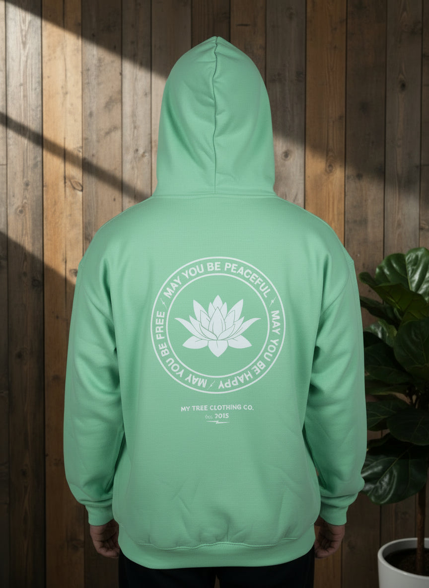 Green hoodie with a white lotus flower design and text may you be peaceful may you be happy may you be free on the back against a wood panel wall and plant.