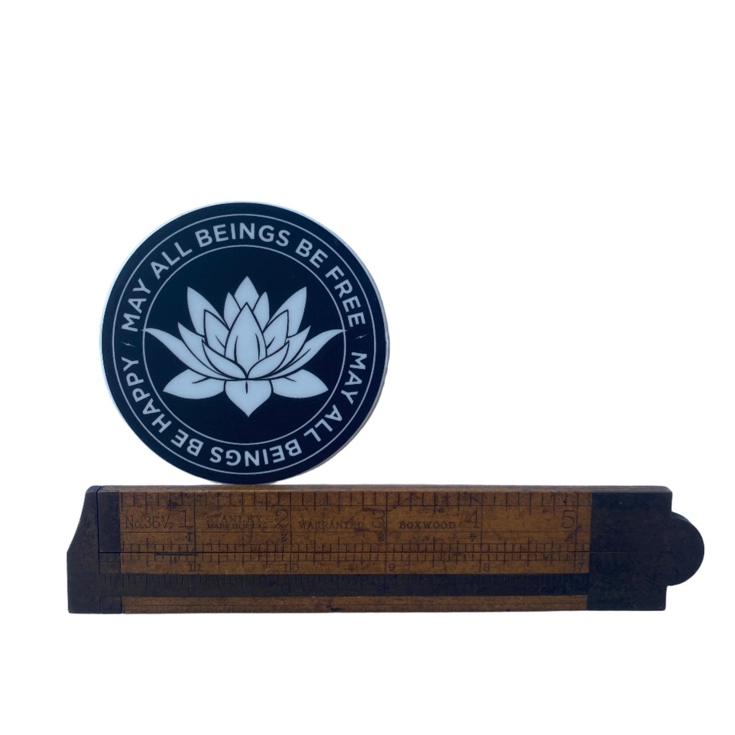 Black circular sticker with white text, may all beings be free-may all beings be happy with lotus flower design next to a ruler on a white background.