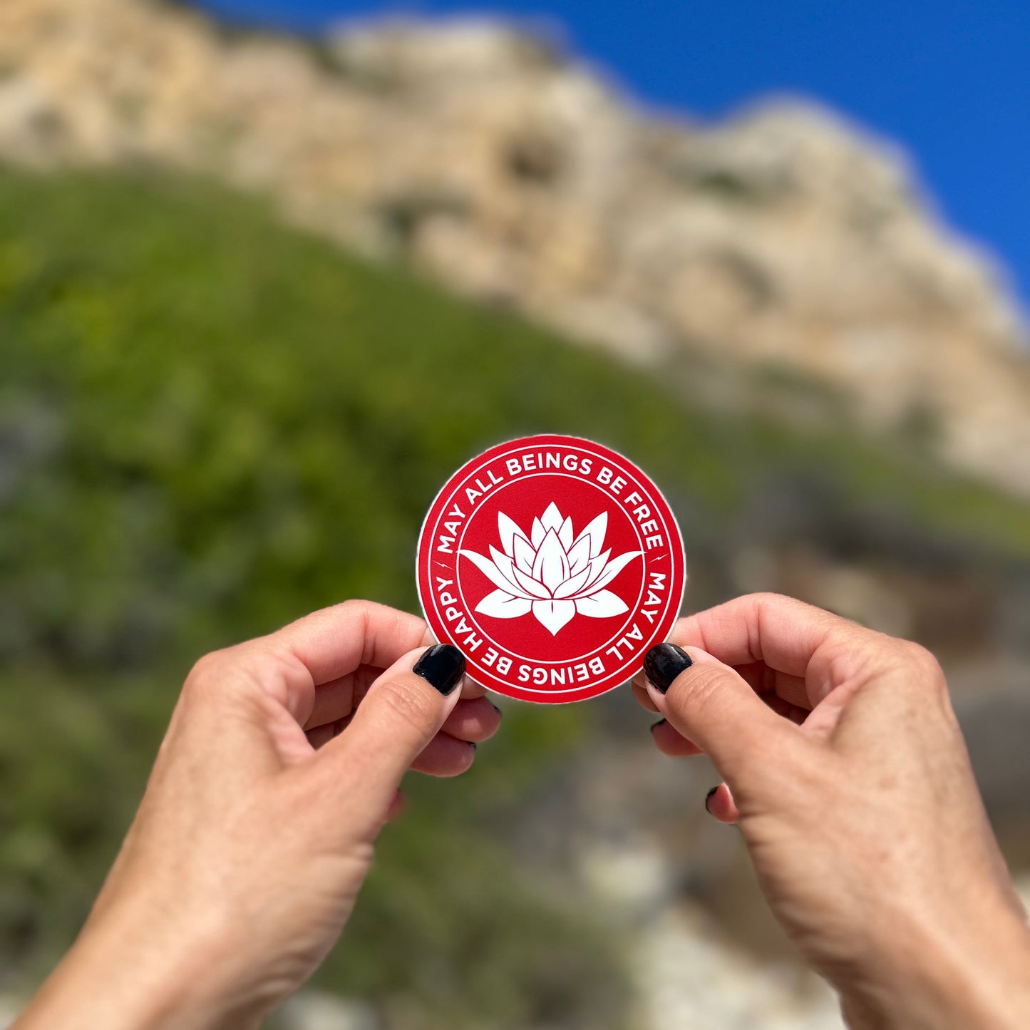 Round red and white three inch sticker being held in a hand with a lotus in the center and the Buddhist prayer, may all beings be happy-may all beings be free. With the blue sky in the background.