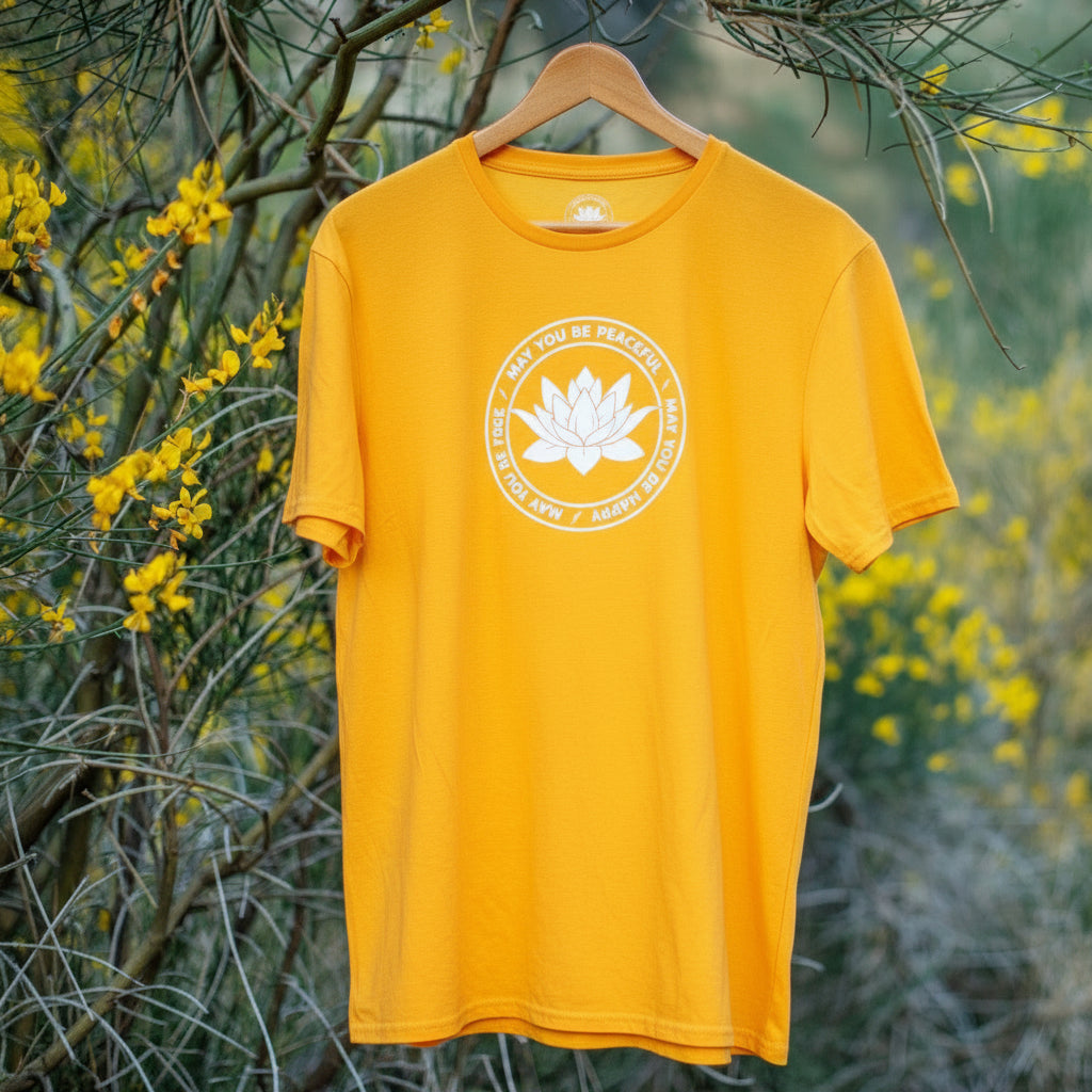 Yellow t-shirt with a white lotus flower graphic, may you be peaceful may you be happy may you be free, on a hanger against a natural background with yellow flowers.